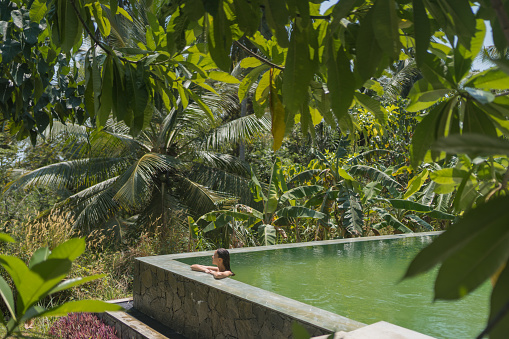 Young woman relaxes on edge of outdoor swimming pool in tropical jungle