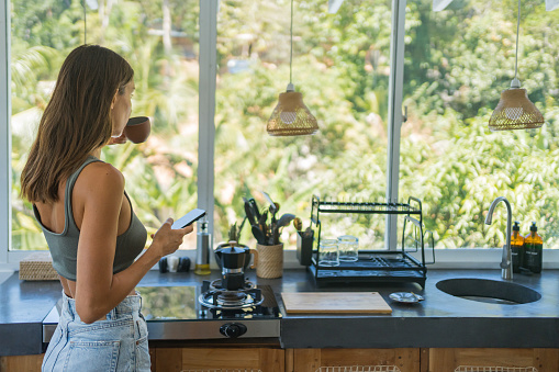 Young woman enjoys morning coffee and uses mobile phone in modern kitchen in rainforest setting