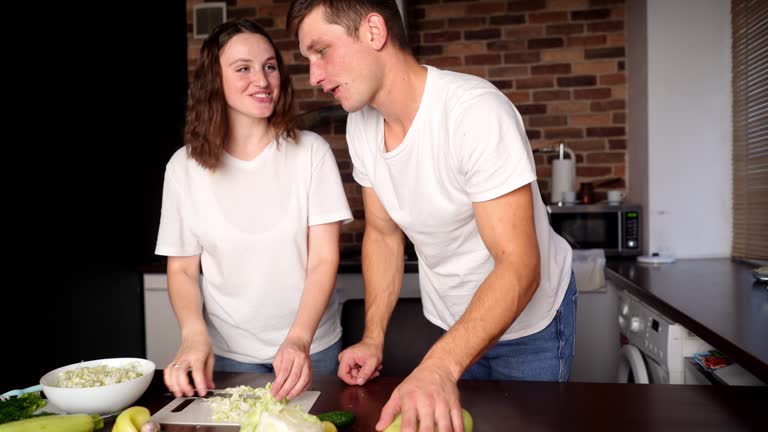 Husband and wife prepare salad together in the kitchen, cut vegetables, woman gives man to try vegetables. Concept of healthy eating, vegetarianism, cooking at home