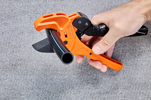 Hand of plumber holds pipeline scissors cutter  with ratchet mechanism ready to cut segment of HDPE  pipe, closeup on gray background.