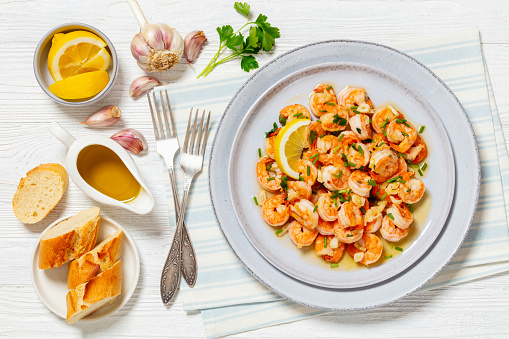 gambas al ajillo, easy spanish garlic shrimp are sauteed in olive oil and lots of garlic served on plate with chives and lemon on white wooden table with crusty bread, flat lay