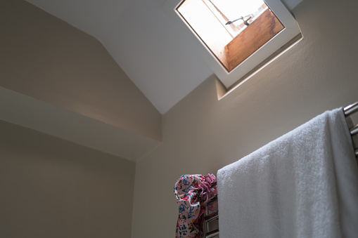 Shallow focus of a damp shower towel and shower cap hanging on a radiator. A small skylight window is opened to help remove the hot steam after taking a shower.
