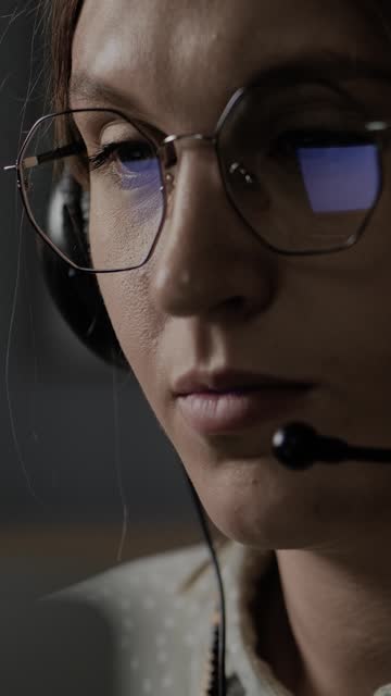 Woman is talking on headset. Attractive woman in glasses talks on headset with microphone, glasses reflect the screen of computer, monitor, laptop