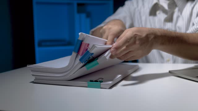 A man is holding a stack of papers with blue and green stickers on them. The stack is on a table and the man is looking at it. He is working overtime because there is a lot of work.