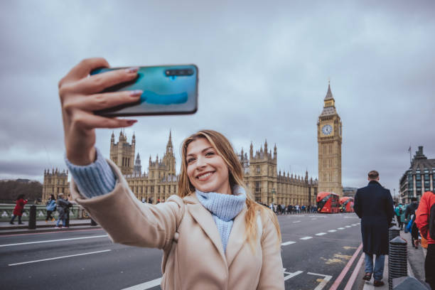 Wide angle panoramic candid portrait of a young cheerful blond woman in her 30s exploring the city and taking selfie with her smart mobile phone device with toothy smile at Westminster bridge in down town capital city of London, England. Selective focus with priority over the model including plenty of copy space on the defocussed background, which consists the iconic architecture of Parliament building and Big Ben clock tower. Photo created during cold season outdoors and the model is with warm casual clothes inc light cream and blue colours on a cloudy day - creative stock photo joven alegre mujer rubia en sus 30 años con la sonrisa de tooty está tomando selfie con su dispositivo de teléfono móvil en el centro de la ciudad cerca de la torre del reloj big ben, ciudad de londres, inglaterra - foto de archivo creativa