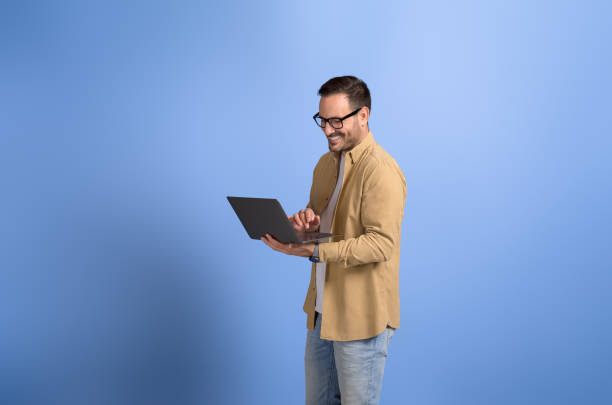 istock Young businessman smiling and working online over laptop while standing on isolated blue background 2088474379