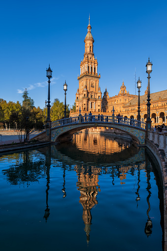 Seville, Andalusia, Spain - October 24, 2023: Plaza de Espana at sunrise, North Tower and bridge over canal with reflection in water, city landmark.