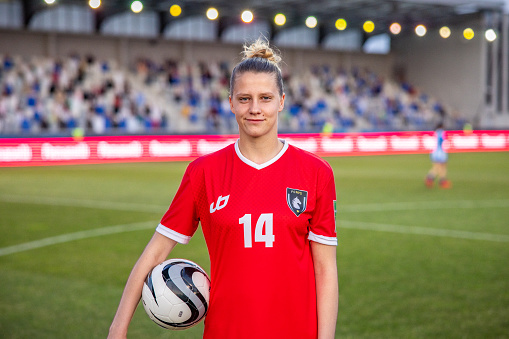 A focused female Caucasian football player in a professional red kit holds a soccer ball at an outdoor stadium. She exhibits a determined stance with the field and spectators in the background.