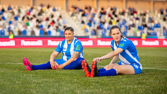Two Caucasian female soccer players in blue and white uniforms stretching on the grassy field. One sitting and one lying down, they are preparing for a football match. The stadium is filled with spectators in the background, indicating a professional sports event outdoors.