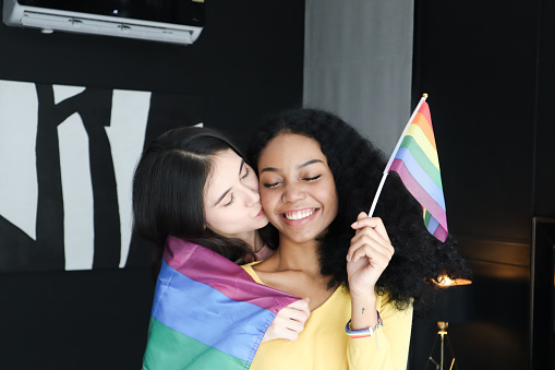 Two ethnically diverse lesbians kissing proudly about their homosexuality during pride month,attractive young women of the same gender holding rainbow flags, human rights and gender equality concepts.