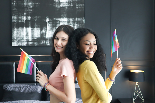 Two ethnically diverse lesbians smiling proudly about their homosexuality during pride month, attractive young women of the same gender holding rainbow flags, human rights and gender equality concepts.
