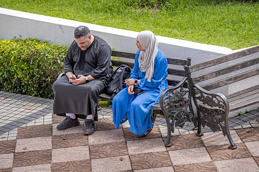 Kuala Lumpur, Malaysia - January 5th 2024: Couple in Islamic dress sitting on a bench outside a public park