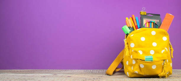 istock Yellow backpack with white polka dots with different colorful stationery on table. Purple background. Back to school. Banner design. 2088656919