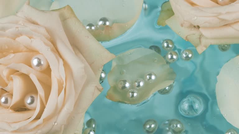 Pearls fall on the surface of the water with petals and flowers of beige roses on a blue background.