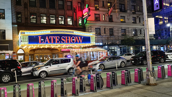 Manhattan, New York, USA - September 21, 2023: Evening shot of the neon sign of Broadway Ed Sullivan Theater. Cars and a cyclist pass by. In the foreground a bicycle storage for rental bicycles