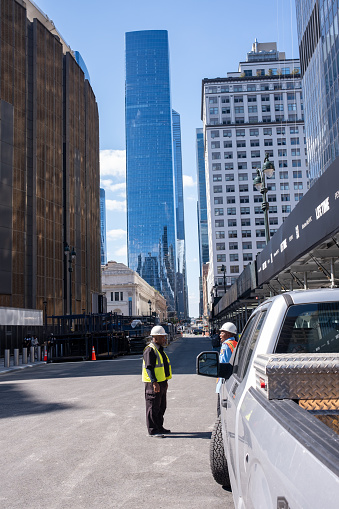 Manhattan, New York, USA - September 20, 2023: Two workmen wearing hard hats talk next to a truck on an empty street next to Madison Square Garden. A skyscraper in the background.