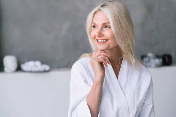 Middle aged woman in white bathrobe looking away, standing in bathroom