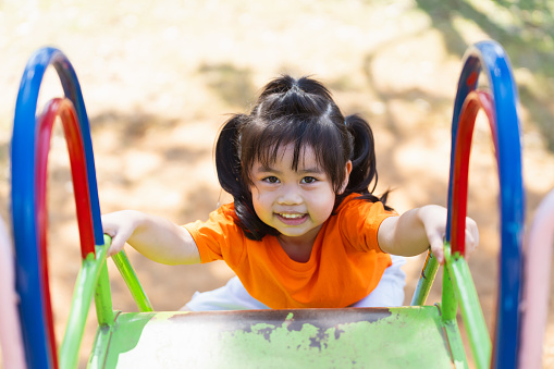 A young girl is smiling as she climbs a green and blue playground slide. The slide is located in a park, and the girl is wearing an orange shirt