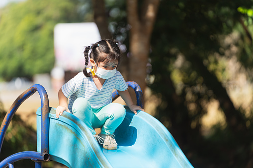 A little girl wearing a mask is sitting on a blue slide. The slide is old and rusty, and the girl is looking up at the sky