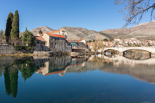 Trebinje's old town reflects in river, early springtime, bridge spans water, cypress trees, mountains behind, clear blue sky. Trebinje, Bosnia and Herzegovina