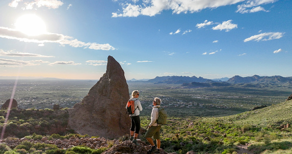 Mature hikers explore rock ridge crest above desert at sunset and look rock pinnacle in distance