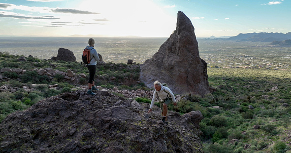 Mature hikers explore rock ridge crest above desert at sunset and look rock pinnacle in distance