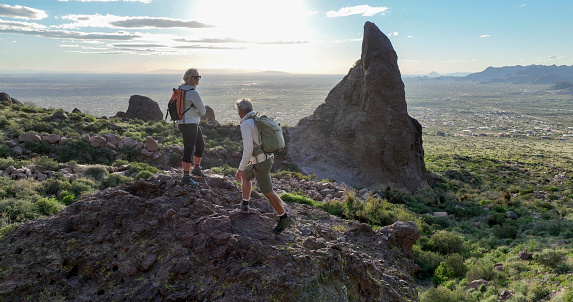 Mature hikers explore rock ridge crest above desert at sunset and look rock pinnacle in distance
