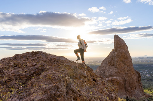 Mature woman ascends rock ridge crest above desert at sunset, rock pinnacle and city in distance