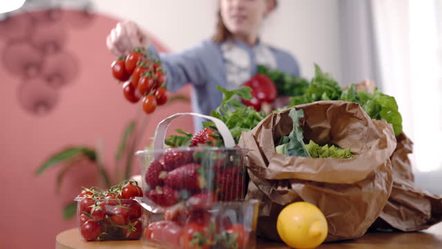 Fresh organic vegetables and fruits in eco paper bags and plastic containers on wooden table in the kitchen, defocused woman pulling out groceries from package and putting on table, process of checking online order, food delivering concept