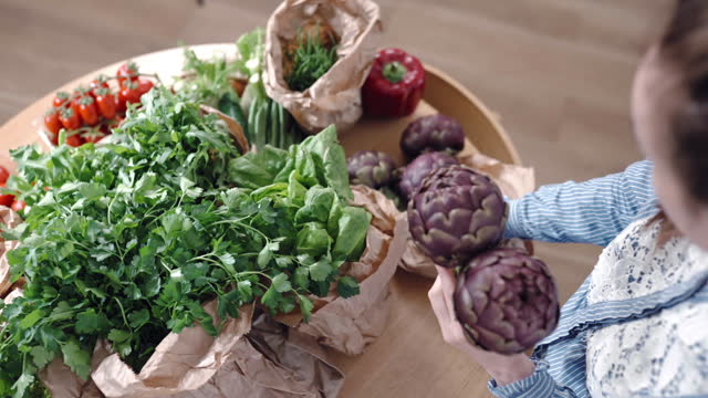 Organic vegetables and fruits being delivered by courier service at home, top view shot of woman pulling ripe artichokes out of paper bag, variety of greenery and healthy vegetables on kitchen table, food online shopping concept