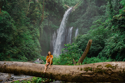 Serene woman crossing river by log on the background of tropical waterfall while hiking