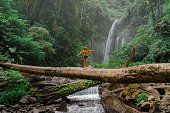 Woman crossing river by log on the background of tropical waterfall while hiking