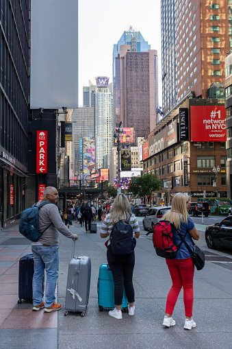 Manhattan, NY, USA - September 20, 2023: A man and two women with travel suitcases stand on a sidewalk next to a busy street. Commercials and skyscrapers can be seen in the background.