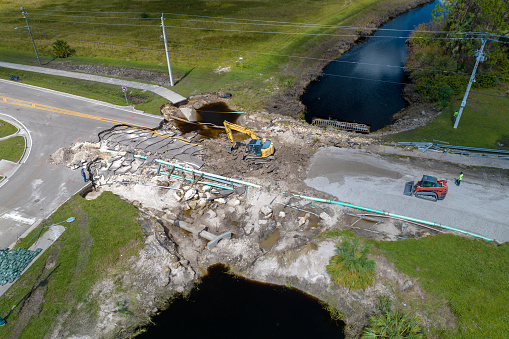 Road construction. Excavator repairing destroyed bridge after hurricane flooding water washed away asphalt in Florida. Construction equipment at roadwork site.