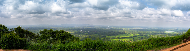 An afternoon panoramic view from the top of Sigiriya Rock, overlooking a vast green forest with distant mountain ranges under a blue sky.