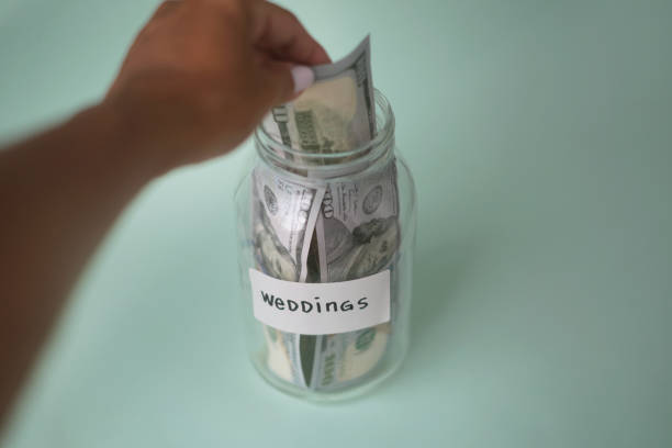 Close-up shot unrecognizable woman inserting US currency money  into her wedding glass jar . concept : saving money in a glass jar.
