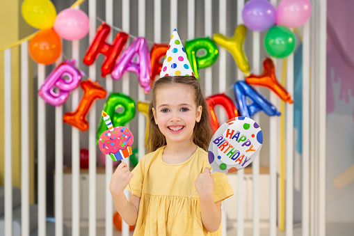 The birthday girl in a festive hat, with a pipe and props is having fun on the background of a multi-colored wall with balloons