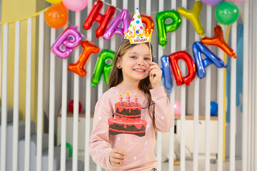 The birthday girl in a festive hat, with a pipe and props is having fun on the background of a multi-colored wall with balloons