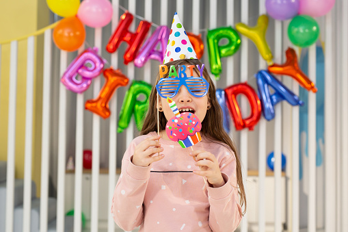 The birthday girl in a festive hat, with a pipe and props is having fun on the background of a multi-colored wall with balloons