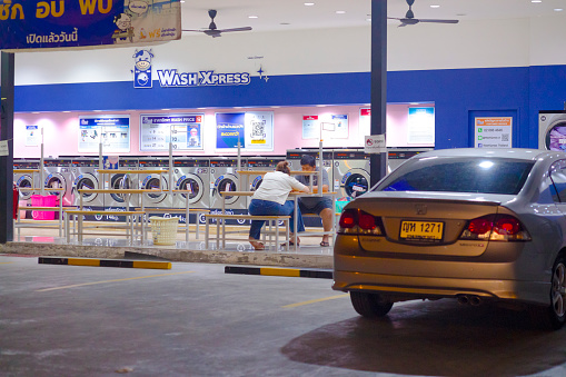 Adult thai couple is sitting at laundromat shop in street Senanikhom at night. People are relaxing and waiting for laundry and waching machine. Both are using mobile phones. At right side a car is parked. People are wearing casual clothing