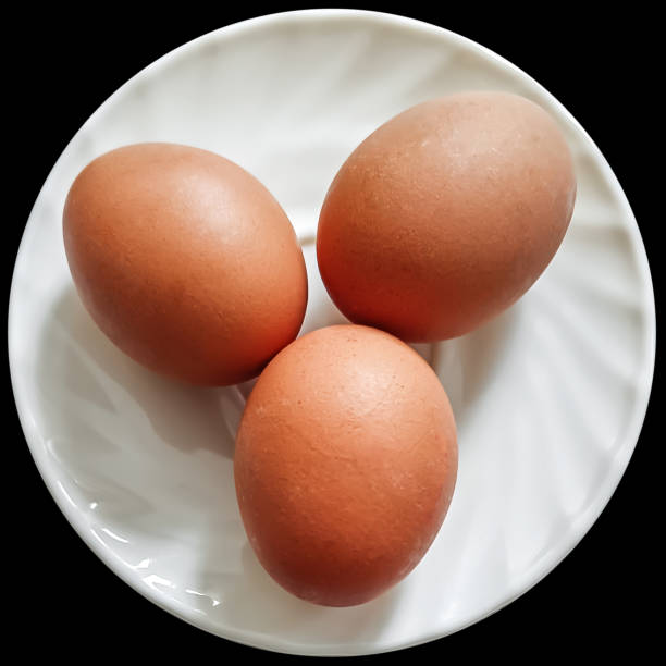 Three fresh chicken eggs on ceramic white plate, isolated on black background, viewed directly above.