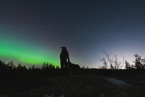 Silhouette of a man with a night vision device on his head and with a dog in nature at night against the backdrop of the starry sky and northern lights. High quality photo