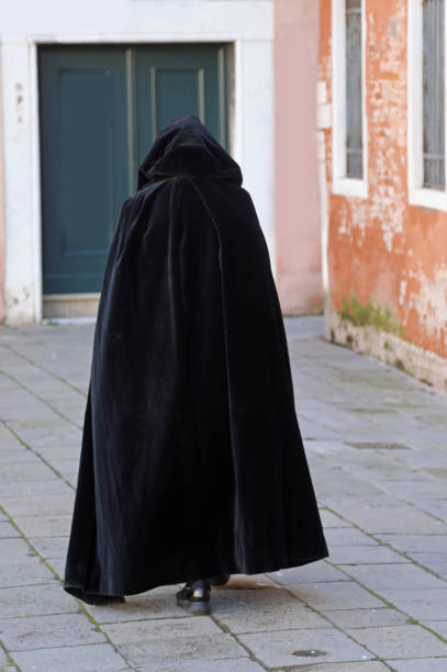 un personnage portant une capuche se promène dans une ruelle étroite de la ville, vêtu d’un tabard noir usé - tabard photos et images de collection