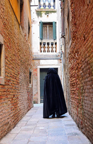 un personnage portant une capuche se promène dans une ruelle étroite de la ville, vêtu d’un tabard noir usé en guise de cape - tabard photos et images de collection