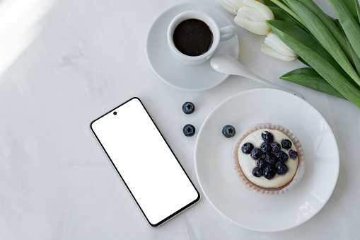 Mobile phone with blank screen mockup, coffee cup, panna cotta dessert and flowers on white table background with natural sunlight, aesthetic business branding template, small business concept.