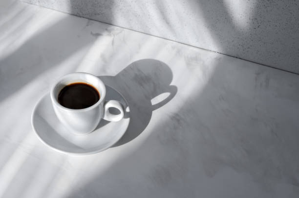 Cup with coffee drink, saucer on neutral white marble table background with abstract natural sunlight shadows, minimal aesthetic good morning concept, elegant business branding backdrop, copy space