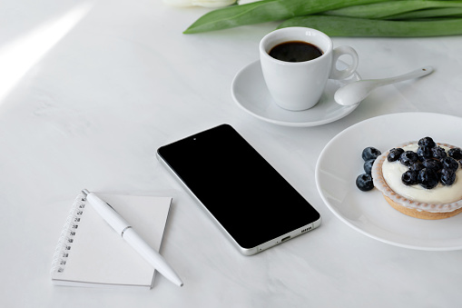 Smartphone and notebook, pen, cup of coffee and panna cotta dessert on white table background with natural sunlight, business morning breakfast, minimal aesthetic home office workspace.