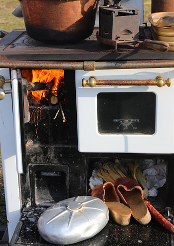 Old wood-fired stove in a farmhouse kitchen with a lit fire and a small pot for heating water and old peasant shoes with a copper cauldron on top for heating