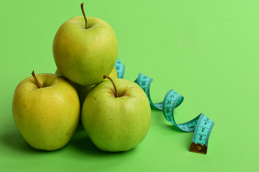 Sports and healthy regime symbols. Diet and sport regime concept. Apples in bright green color and twisted measure tape on green background. Tape measure in cyan color near juicy fruit, close up