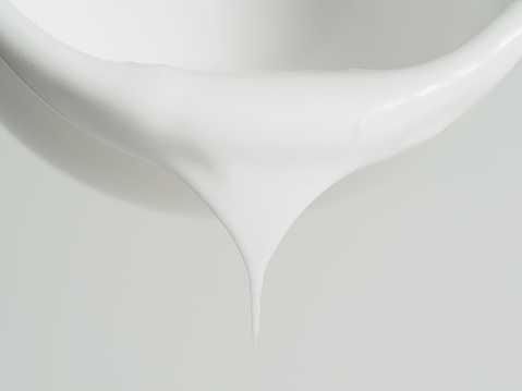 A poised drip of white paint elegantly extends from the smooth edge of a ceramic bowl against a soft white backdrop.